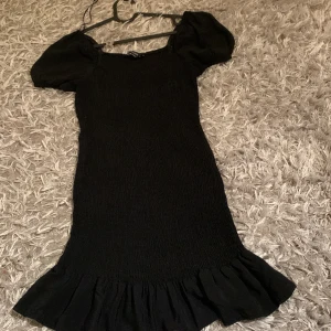 Svart volangklänning med puffärm - Supersöt svart klänning med puffärmar och volang nertill. Smockad överdel ger en tight passform och klänningen har en feminin siluett. Perfekt för dig som vill ha en enkel men trendig look.