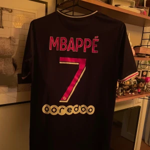 PSG fotbollströja Mbappé #7 - Säljer en officiell Paris Saint-Germain fotbollströja från Nike med Mbappé och nummer 7 på ryggen. Tröjan är mörkblå med röda och vita ränder, kortärmad och har klubbmärke samt sponsortryck på bröstet. Perfekt för fans av PSG och Mbappé.