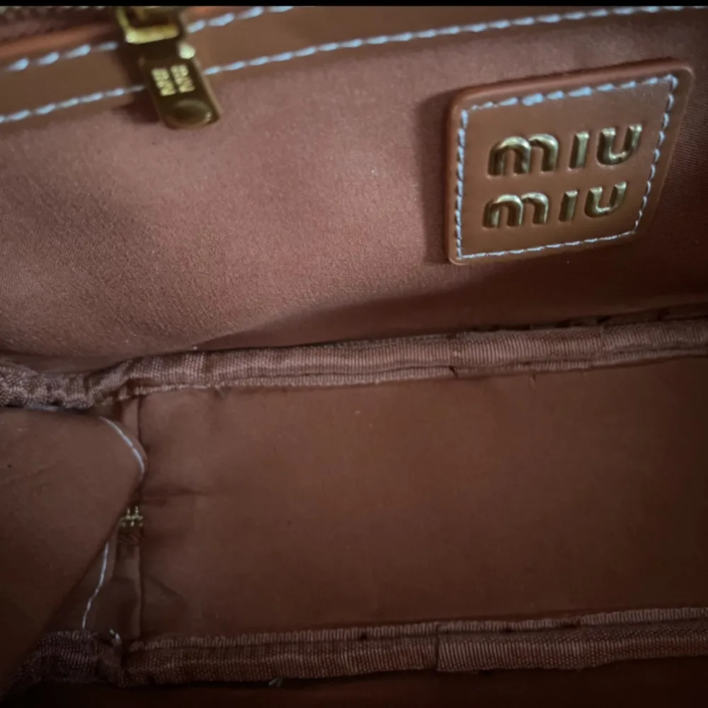 Snygg beige handväska från Miu Miu med bruna detaljer och logga framtill. Väskan har dragkedja upptill och är tillverkad i textil med inslag av läder. Perfekt för dig som vill ha en stilren accessoar med lyxig känsla. Ingen kvitto eller köpbevis finns för att bevisa äktheten. Laukut & Käsilaukut.