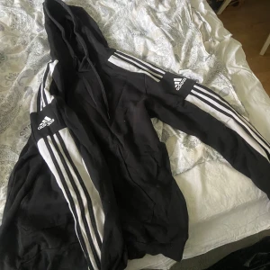 Svart Adidas hoodie med vita ränder - Svart hoodie från Adidas med klassiska vita ränder längs ärmarna och Adidas-logga på bröstet och ärmen. Modellen har huva och dragkedja framtill. Perfekt för en sportig stil.