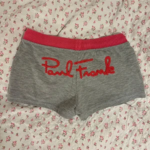 Grå shorts från Paul Frank med rosa detaljer - Grå mjuka shorts från Paul Frank med bred rosa resår i midjan och rosa snörning. Shortsens baksida har ett stort rött Paul Frank-tryck och framsidan har två fickor med rosa kant samt ett litet apmärke och hjärta.