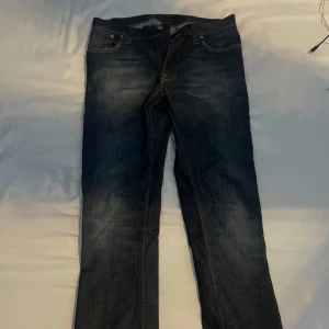 Mörkblå jeans från Nudie Jeans - Säljer ett par mörkblå jeans från Nudie Jeans med klassiska kontrastsömmar och broderade bakfickor. Jeansen har normal passform och raka ben, perfekt för en stilren vardagslook.
