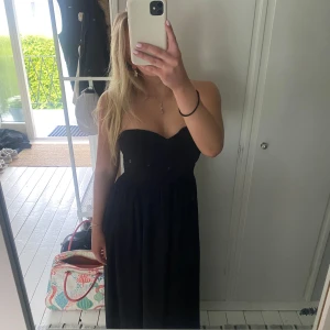 Svart axelbandslös klänning - Säljer en svart axelbandslös balklänning med omlott-detalj över bysten och markerad midja. Klänningen har en mjukt fallande kjol och är perfekt för bal eller annan tillstllning. Enkel och stilren design som passar till många olika looks.