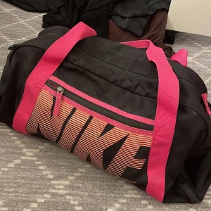 Svart och rosa träningsväska från Nike - Stor svart träningsväska från Nike med breda rosa axelband och detaljer. Väskan har ett stort huvudfack, dragkedja och ett fack framtill med Nike-logga i rosa och orange. Perfekt för gymmet eller sportaktiviteter.