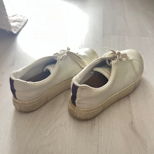 Vita sneakers eytys - Säljer ett par vita sneakers med snörning och lila detalj på hälen. Skorna har rund tå och platt sula, perfekta för en avslappnad stil. Klassisk design som passar till det mesta. Slitna men mest smutsiga, de går att få fräscha!