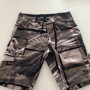 Mönstrade badshorts med fickor - Säljer ett par badshorts med svart och grått grafiskt mönster och snörning i midjan. Shortsen har flera fickor, bland annat en stor benficka med lock. Perfekta för stranden eller poolen.