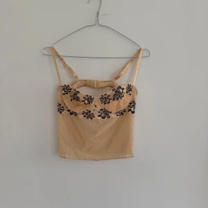 Beige topp/bh i spets - Säljer en beige linnetopp/bh med svarta blommiga spetsdetaljer upptill. Toppen har tunna axelband och en öppen rygg. Perfekt för festen!