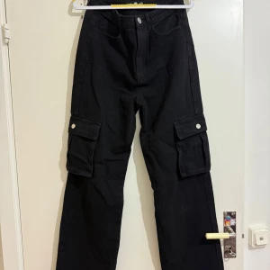 Svarta cargobyxor från Boohoo  - Svarta cargobyxor från Boohoo Blue med raka ben och stora fickor på sidorna med lock och knapp. Byxorna har normal passform och är tillverkade i ett kraftigare bomullsmaterial. Perfekta för en avslappnad och trendig stil.