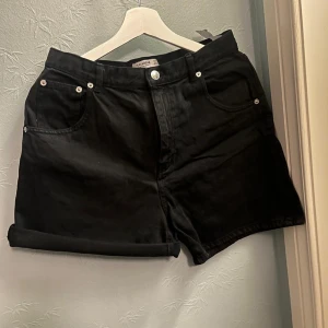 Svarta jeansshorts från Perfect Jeans - Svarta jeansshorts med klassisk femficksdesign och uppvikta benslut. Stilren look med silverfärgade nitar och knapp. Perfekta för en avslappnad stil under varma dagar.