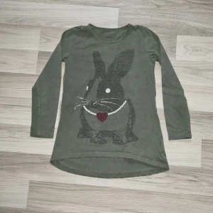 Olivgrön långärmad tröja med kaninmotiv från Lindex  - Säljer en olivgrön långärmad tröja från Lindex med ett stort kanintryck framtill. Kaninen har ett halsband med hjärta och pärlor. Tröjan har rund halsringning och är lite längre baktill. Perfekt för dig som gillar gulliga motiv!