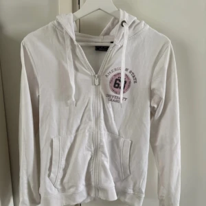 Vit hoodie med dragkedja från OTP Leisurewear - Vit hoodie med dragkedja från OTP Leisurewear. Tröjan har huva, fickor framtill och tryck med text och siffror på bröstet. Perfekt för en avslappnad stil. Använd endast en gång, legat ett tag i garderob därav lossnat lite på dragkedja. Strl S