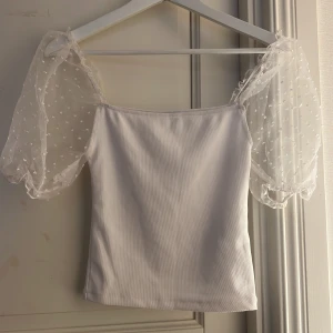 Vit topp med puffärmar i mesh - Säljer en söt vit topp med transparenta puffärmar i mesh och små broderade prickar. Toppen har en rak passform och är perfekt för sommaren