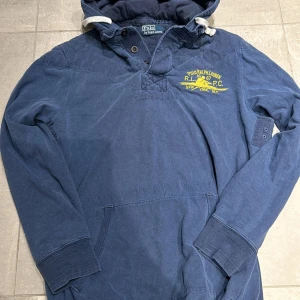 Ralph Lauren Hoodie - Tja! Säljer denna snygga Ralph Lauren hoodien. Den är som en pike men med en huva. Snygg marinblå färg med ett gult tryck. Storlek Medium men kan passa lite större. Hör av vid frågor!
