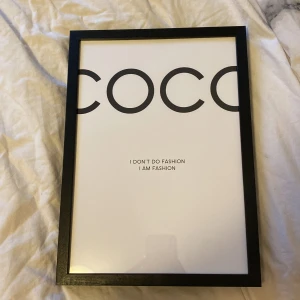 Tavla med texten COCO - Snygg tavla med svart ram och vit bakgrund. Texten 'COCO' i stora svarta bokstäver upptill och citatet 'I DON'T DO FASHION I AM FASHION' i mindre text i mitten. Perfekt som stilren inredningsdetalj. KÖPT FRÅN DESENIO