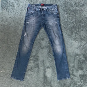 Blå slitna jeans från Jack & Jones - Säljer ett par blå jeans från Jack & Jones med slitningar och lätt tvättad look. Storlek 33/32. Byxorna har fem fickor, dragkedja och knapp i midjan. Skinny/slim passform och normal midja. Perfekta för en avslappnad stil.