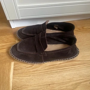 Bruna mockaloafers med kontrastsöm - Säljer ett par bruna loafers i mocka med dekorativ kontrastsöm runt sulan. Skorna har en klassisk design med rund tå och platt sula, perfekta för en avslappnad men stilren look.