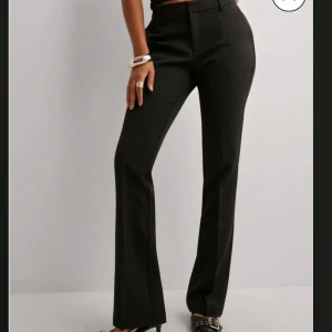 Svarta kostymbyxor med bootcut - Säljer ett par svarta kostymbyxor med bootcut passform och låg midja från Neo Noir i storlek S. Byxorna har klassisk design med hällor för bälte och stängs med dragkedja och knapp framtill. Perfekta för en stilren look.