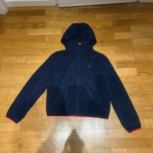 Mörkblå vindjacka från Polo Ralph Lauren - Säljer en mörkblå vindjacka från Polo Ralph Lauren med huva och dragkedja. Jackan har röda detaljer vid ärmslut och nederkant samt en liten röd logga på bröstet. Perfekt för blåsiga dagar.