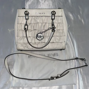 Vit axelväska med volang från Michael Kors. Äkta  - Säljer en snygg vit axelväska från Michael Kors med volangdetaljer och silverfärgade metalldetaljer. Väskan har både handtag och en avtagbar axelrem samt MK-logga framtill. Perfekt accessoar för att lyfta din outfit.Skydds påsen kommer med.