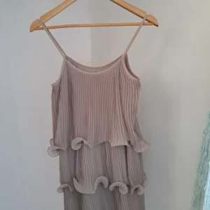 Beige plisserad klänning med volang från H&M - Säljer en beige plisserad klänning från H&M med smala axelband och volangdetaljer. Klänningen har en luftig passform och är perfekt för varma dagar. Materialet är lätt och ger ett fint fall.