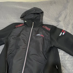 Svart vindjacka Tommy Jeans L - Svart vindjacka från Tommy Jeans med huva och dragkedja. Jackan har broderade loggor på bröstet och ärmen, samt mesh-detaljer på axlarna. Materialet är en mix av polyester och syntet, perfekt för blåsiga dagar. Snygg och sportig look med klassiska Tommy-färger i detaljerna.