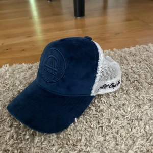 Sprillansny blå och vit keps från All Cap One - Snygg keps i blått och vitt från All Cap One. Framsidan är i blått tyg med broderad logotyp, medan baksidan är i vit med justerbar knäppning. Cepsen är one size så passar dörför på alla.