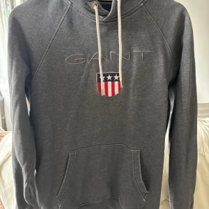 Grå hoodie från GANT - Säljer en grå hoodie från GANT med broderad logga och amerikansk sköld på bröstet. Tröjan har huva med vita snören och en stor magficka framtill. Perfekt för en avslappnad stil.