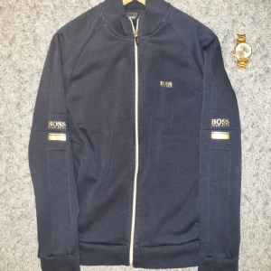 BOSS - Snygg mörkblå jacka från BOSS med vit dragkedja och logga på bröstet samt ärmarna. Jackan har lång ärm och ribbade muddar. Perfekt för en stilren och sportig look.