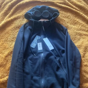 Svart hoodiejacka från C.P. Company - Svart hoodiejacka från C.P. Company med ikoniska goggles i huvan. Jackan har dragkedja framtill, två fickor med dragkedja och justerbar huva med snörning. Tillverkad i mjukt bomullsmaterial för en clean och modern look.