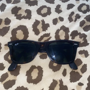 Ray-Ban Wayfarer svarta solglasögon - Klassiska Ray-Ban Wayfarer solglasögon i svart med mörka glas. Snygg, tidlös design med bred båge och diskret logga på sidan. Handgjorda i Italien och perfekta för att lyfta vilken outfit som helst.