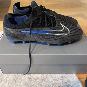 Nike Phantom Gx FG Elite fotbollsskor svart/blå - Nike Phantom Gx FG Elite fotbollsskor i svart med blå och vita detaljer. Ovandel i stickad syntet med mönstrad struktur för bättre bollkontroll. Skorna har snörning och blå yttersula med fasta dobbar för gräsplan. Snygg, modern design för dig som vill sticka ut på planen.