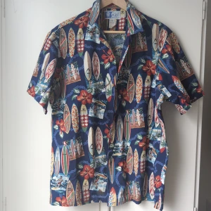 Blå Hawaii-skjorta från RJC, L - Färgstark Hawaii-skjorta från RJC med surfbrädor, blommor och strandmotiv på blå botten. Skjortan har korta ärmar, klassisk krage och knäppning framtill. Perfekt för sommarens chillaste dagar. Tillverkad i mjuk bomull.