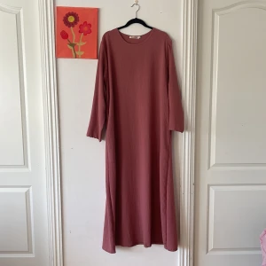 Rosa långklänning från Etoile D'orient med fickor - Långklänning i en dämpad rosa nyans från Etoile D'orient. Klänningen har lång ärm, rund halsringning och är tillverkad i ett lätt, strukturerat tyg. Enkel och flowy passform som ger en chill vibe. Perfekt för dig som gillar modest fashion. 