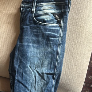 Replay blå jeans med slitningar - Snygga blå jeans från Replay med coola slitningar och tvättade detaljer. Klassisk femficksmodell med tydliga kontrastsömmar och Replay-logga på fickan. Jeansen har en modern passform och är perfekta för en avslappnad streetstil.