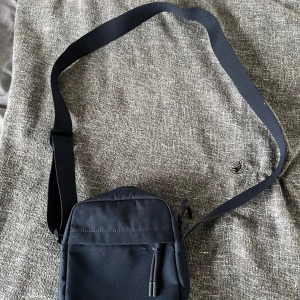 Svart axelväska från Lacoste - Säljer en stilren blå  axelväska från Lacoste med klassisk krokodillogga framtill. Väskan har justerbar rem, dragkedja och är tillverkad i slitstark polyester. Perfekt storlek för mobil, plånbok och andra småsaker. Enkel och snygg design som funkar till allt.