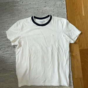 Vit t-shirt från Lacoste med svart krage - Vit t-shirt från Lacoste i bomull med klassisk krokodil-logga broderad på bröstet. T-shirten har en svart ribbad rund halsringning som ger en snygg kontrast. Regular fit och kortärmad modell, perfekt för en clean och stilren look.