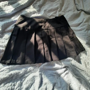 Svart plisserad kortkjol - Snygg svart kortkjol med plisserade veck och bred linning. Perfekt för en trendig och stilren look. Kjolen är enkel att matcha och ger en cool vibe till outfiten.