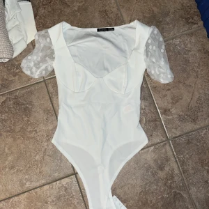 Vit bodysuit med puffärm från Boohoo - Supersnygg vit bodysuit från Boohoo med transparenta puffärmar i mesh och stora prickar. Toppen har markerade kupor och en figurnära passform. Perfekt för dig som vill sticka ut med coola detaljer och trendig look.