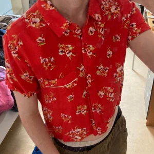 Röd blommig skjorta från Forever 21 - Kortärmad röd croppad skjorta från Forever 21 med blommigt mönster i vitt, rosa och gult. Skjortan har klassisk krage, knappar framtill och en bröstficka med vit kant. Strl S men passar även XS