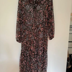 Blommig långklänning från KappAhl - Supersöt långklänning från KappAhl i svart med små rosa och lila blommor. Klänningen har långa ärmar, v-ringning, transparent polyestertyg.  Passar mig som är M-L. Då klänningen något transparent ingår en kort svart klänning i stl S från Indiska att ha under.