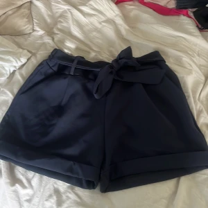 Mörkblå shorts med knytband - Snygga mörkblå shorts med hög midja och knytband i midjan. Shortsen har uppvikta benslut och är tillverkade i ett mjukt, lite glansigt material. Perfekta för sommardagar och enkel att matcha med olika toppar.