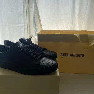 Svarta sneakers från Axel Arigato med krokodilpräglat mönster - Säljer ett par svarta sneakers från Axel Arigato med snygg krokodilpräglad yta. Skorna har snörning och rund tå, och kommer med originalkartong och dustbag. Perfekta för dig som gillar stilrena och exklusiva sneakers. Skorna är också nästan helt nya 