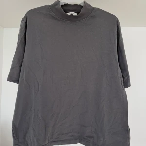 Mörkgrå t-shirt med hög krage - En enkel och stilren mörkgrå t-shirt med högre krage. T-shirten har korta ärmar och en rak passform, perfekt för en avslappnad look. Passar bra till jeans eller shorts.