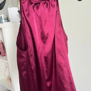 Vinröd ärmlös halterneck topp i satin - Säljer en vinröd ärmlös halterneck topp i glansigt satinmaterial.  har en lös passform och en liten öppning med knäppning i nacken. Perfekt för dig som gillar en elegant och enkel stil.