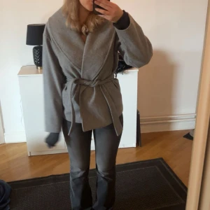 Grå omlottjacka med bälte - Snygg grå jacka i omlottmodell med bälte i midjan. Jackan har lång ärm och en stilren, enkel design utan synliga knappar eller dragkedja. Perfekt för dig som gillar en clean och modern look. Använd någon få gång