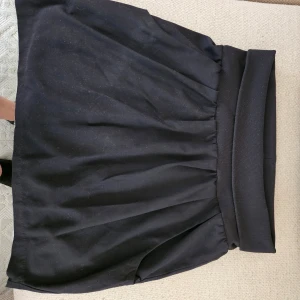 Svart kortkjol med hög midja - Säljer en svart kortkjol med hög, bred midja och lätt utställd form. Kjolen har en enkel och stilren design som passar till många olika outfits. Perfekt för dig som gillar en klassisk look.