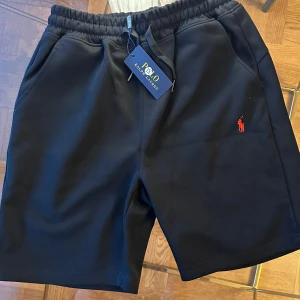 Svarta shorts från Polo Ralph Lauren - Svarta shorts från Polo Ralph Lauren med elastisk midja och snörning. Klassisk röd broderad logga på vänster ben. Mjukt material och två sidofickor. Perfekta för en avslappnad stil.