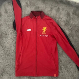 Liverpool FC träningsjacka från New Balance - Röd träningsjacka från New Balance med Liverpool FC-logga och broderad text för 125-årsjubileum. Jackan har svarta detaljer längs ärmarna, dragkedja framtill och hög krage. Perfekt för fotbollsfans som vill visa sitt stöd för LFC.