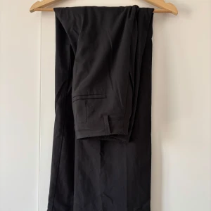 Svarta kostymbyxor - Klassiska svarta kostymbyxor med raka ben. Byxorna har hällor för bälte och stängs med dragkedja och knapp. Perfekta för en stilren look. Storleken säger XXS men jag skulle säga att de är en normal S. Bra men använt skick!