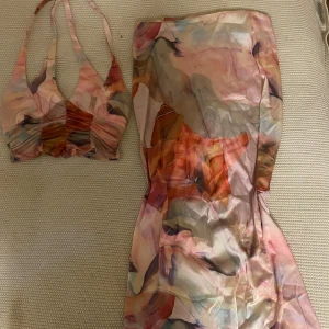 Pastellfärgat set med halterneck - Säljer en långklänning i pastelliga toner med abstrakt mönster. Klänningen har halterneck-top och öppen rygg, och är gjord i ett tunt, glansigt material som känns som satin eller polyester. Färgerna går i rosa, beige, lila och orange. Perfekt för sommarens alla tillfällen! Aldrig använt. 